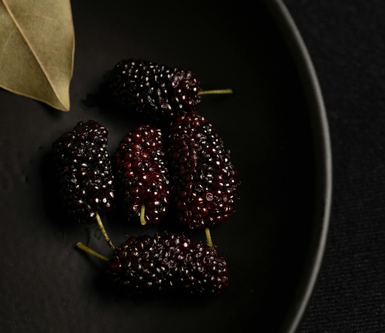 Detailed photo of fresh black mulberries placed on a dark plate with leaf decoration.