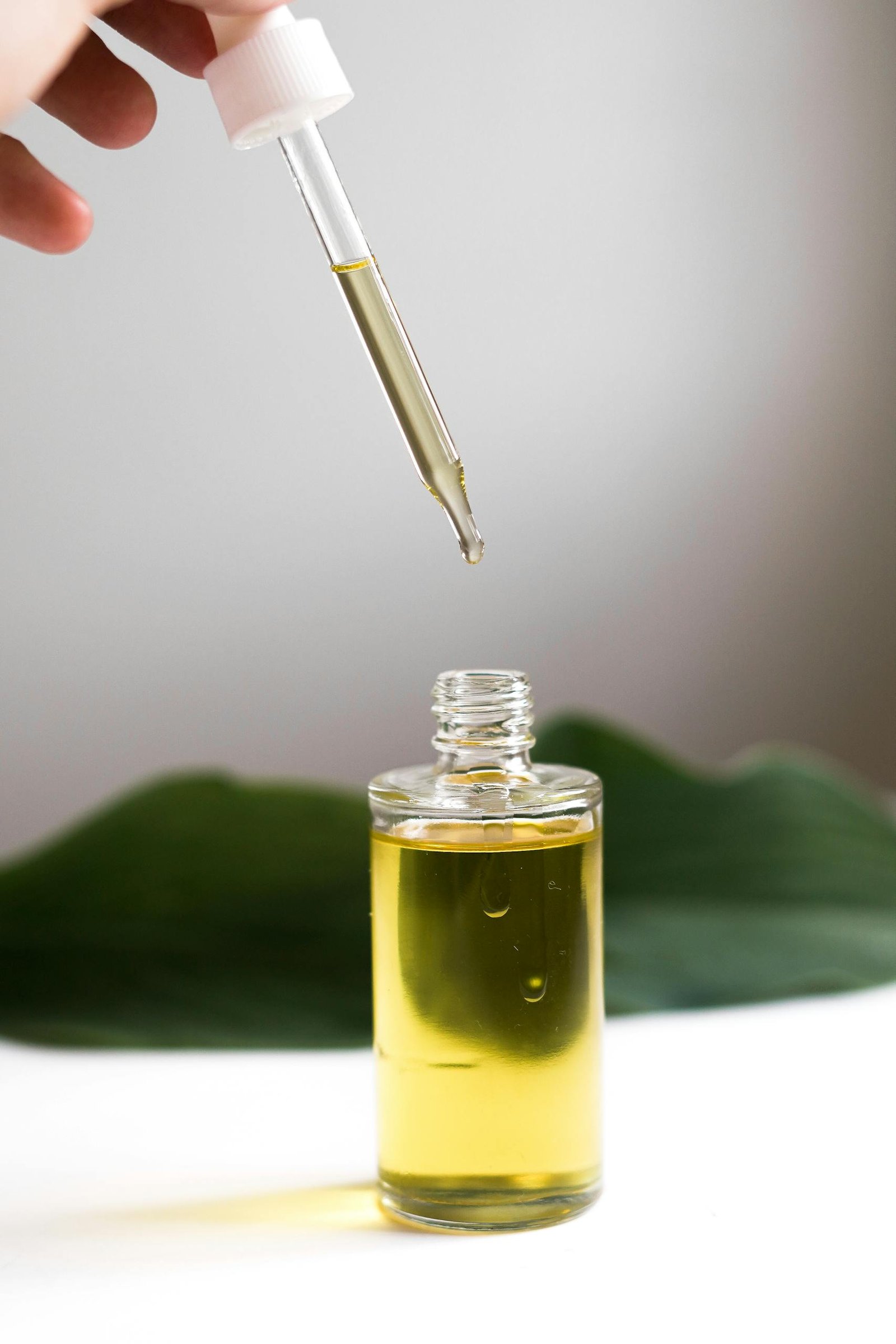 A hand holding a dropper over a bottle of essential oil, with a green leaf background.